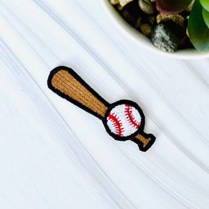 May include: Embroidered iron-on patch of a baseball and bat. The baseball is white with red stitching and the bat is brown with black stitching.