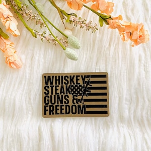 May include: A brown rectangular sign with the text "Whiskey, Steak, Guns & Freedom" in black lettering. The sign features a stylized American flag and a rifle graphic.