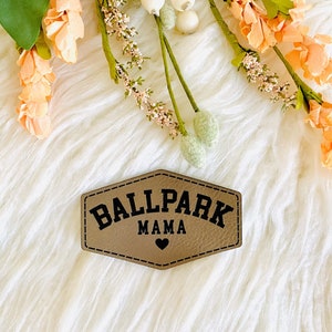 Peut inclure: Un patch en cuir marron avec le texte "Ballpark Mama" en lettres noires et un petit symbole de cœur.