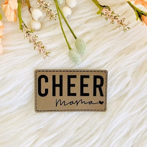 Peut inclure: Un patch en cuir marron avec le texte "CHEER Mama" en lettres noires. Le patch a une bordure cousue et un petit symbole de cœur.