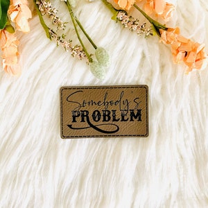 May include: A brown leather patch with the text "Somebody's PROBLEM" in black lettering. The patch is on a white fluffy surface with pink flowers.