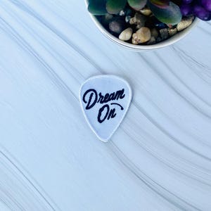 May include: White guitar pick shaped patch with black embroidery that says "Dream On".
