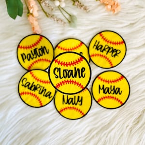 May include: A collection of personalized softball patches. Each patch is yellow with red stitching, resembling a softball. The patches feature names in black script: Payton, Sabrina, Naly, Harper, Maya, and Sloane.
