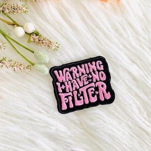 Op de afbeelding: Een geborduurde zwarte en roze patch met de tekst "Warning I Have No Filter" in een retro-lettertype.