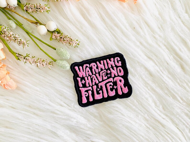Op de afbeelding: Een geborduurde zwarte en roze patch met de tekst "Warning I Have No Filter" in een retro-lettertype.