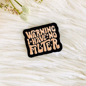 Op de afbeelding: Een zwarte patch met witte stiksels met de tekst "WARNING I HAVE NO FILTER" in een gestileerd lettertype.