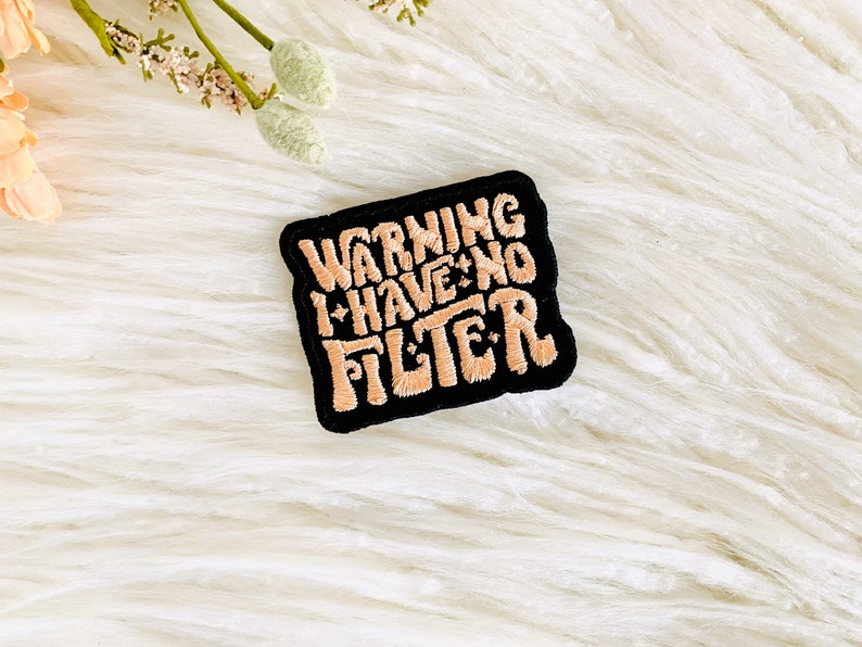 Op de afbeelding: Een zwarte patch met witte stiksels met de tekst "WARNING I HAVE NO FILTER" in een gestileerd lettertype.