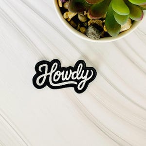 May include: A black and white embroidered patch with the word "Howdy" in a cursive font. The patch has a black border and white lettering. The patch is on a white, textured surface, with a small succulent plant in the background.