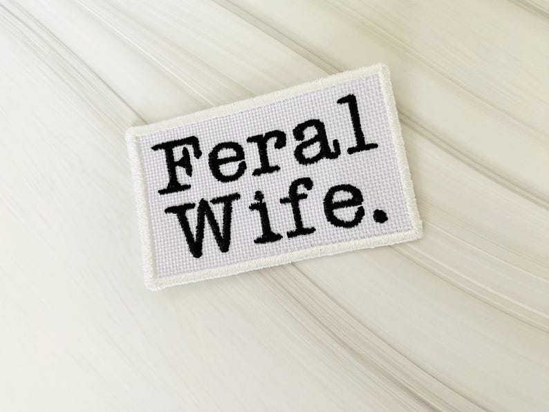 May include: A white rectangular patch with the words "Feral Wife." in bold, black, typewriter-style font. The patch has a white border and is set against a light, textured background.