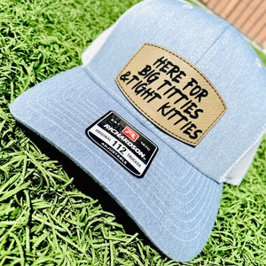 May include: A light blue trucker hat with a white mesh back. The hat has a brown leather patch with the text "HERE FOR BIG TITTIES & TIGHT KITTIES" in black lettering. The hat also has a red and white Richardson label with the text "ORIGINAL 112 TRUCKER ADJUSTABLE".