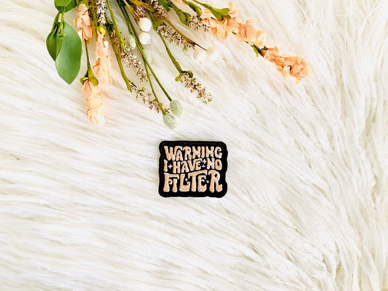 Op de afbeelding: Een geborduurde patch in zwart en wit met de tekst "WARNING I HAVE NO FILTER" in roze.