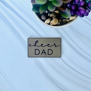 May include: A brown leather patch with the text "cheer DAD" in black lettering.