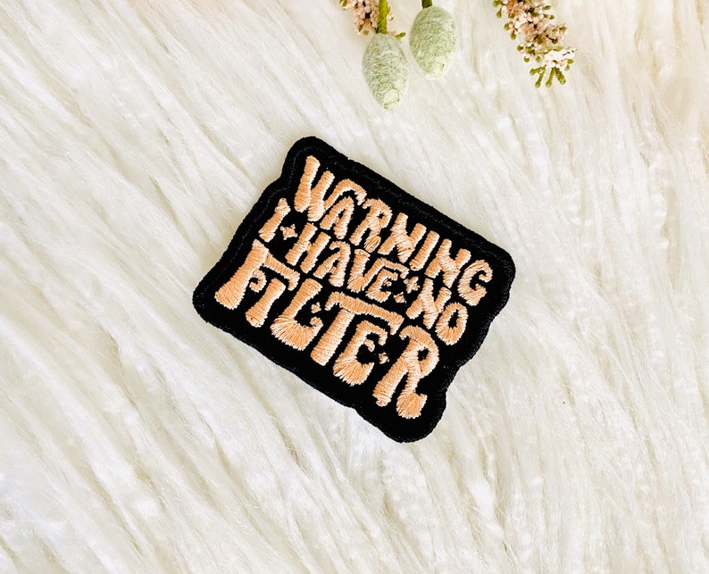 Op de afbeelding: Een zwarte geborduurde patch met de tekst "WARNING I HAVE NO FILTER" in perzikkleurige draad.