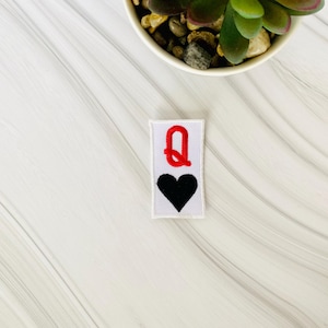May include: White felt patch with red embroidered letter Q and a black embroidered heart. The patch is for sewing or ironing onto fabric.