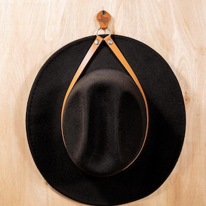 May include: A black hat suspended from a leather strap on a wooden wall. The strap is fastened to the hat with metal rivets. A small leather tag shaped like Texas is fixed to the wall.