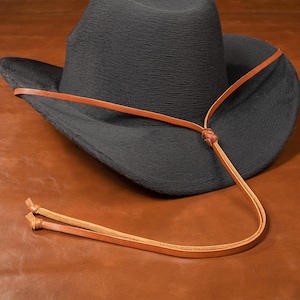 Könnte beinhalten: Ein schwarzer Cowboyhut mit einem braunen Lederband und einem Kinnriemen. Das Band ist hinten am Hut zu einem Knoten gebunden.