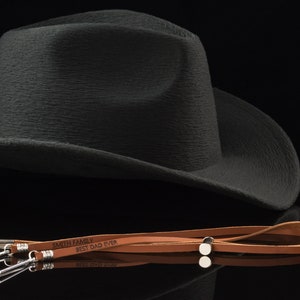 Puede incluir: Un cordón de cuero marrón con herrajes plateados y el texto "Smith Family Best Dad Ever" grabado en él. El cordón está sobre una superficie negra junto a un sombrero de vaquero negro.
