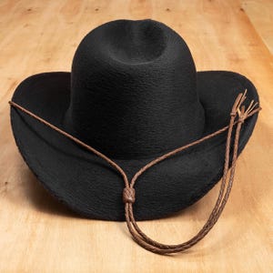 Peut inclure: Un chapeau de cowboy noir avec une surface texturée et une mentonnière en cuir marron. Le chapeau a un large bord et une couronne arrondie. La mentonnière est tressée et nouée au centre.