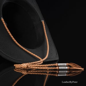 Può includere: Un cappello da cowboy nero con una fascia in pelle marrone chiaro e nappe. La fascia è intrecciata e presenta due inserti in metallo argentato con il nome "MARK" inciso. Il cappello è posto su uno sfondo nero.