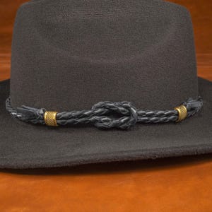 Könnte beinhalten: Ein schwarzer Filzhut im Fedora-Stil mit einem geflochtenen Lederband und goldenen Metallverzierungen.