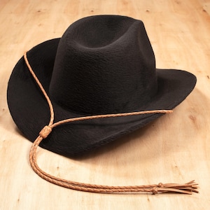 May include: A black cowboy hat with a textured surface and a brown leather chin strap. The hat has a wide brim and a rounded crown. The leather strap is braided and knotted.