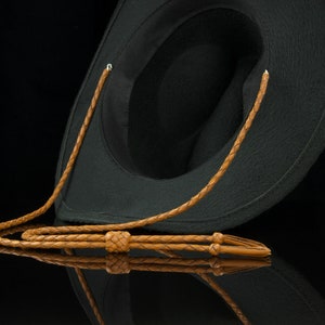 May include: A black cowboy hat with a brown leather chin strap. The hat is made of a felt-like material and has a classic western design. The chin strap is braided and features a sliding knot for adjustment.