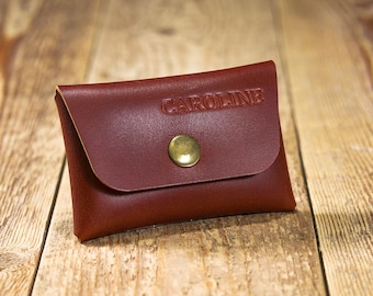 May include: A rectangular brown leather card holder featuring a flap closure secured by a single brass-colored metal snap button. The name CAROLINE is embossed in capital letters on the front flap of the leather accessory.