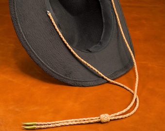 May include: A dark grey hat with a brown leather chin strap. The hat has a soft, textured surface and a wide brim. The chin strap is braided and features brass-coloured tips.