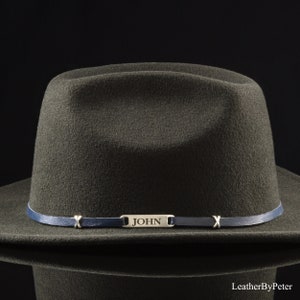 Könnte beinhalten: Ein dunkelgrüner Fedora-Hut mit einem blauen Lederband und einem silbernen Namensschild mit der Aufschrift "JOHN".