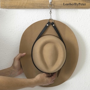 Può includere: Un cappello fedora color cammello sospeso da un appendiabiti in pelle nera. L'appendiabiti è attaccato a una barra di legno con un fermaglio in metallo color argento. Il testo "LeatherByPeter" è visibile.