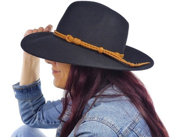 May include: A black fedora hat with a wide brim and a tan leather band. The hat is worn by a person wearing a denim jacket. The hat's design is a classic style, suitable for various occasions.
