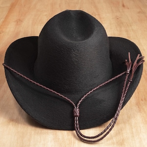 May include: A black cowboy hat with a textured, felt-like surface and a pinched crown. The hat features a dark brown braided leather cord hatband that wraps around the base, with two long, dangling braided ends hanging down the side.