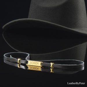Puede incluir: Una pulsera de cuero negro con una placa dorada grabada con el nombre "JOHN". La pulsera tiene un diseño de doble correa y detalles dorados. Un sombrero fedora gris oscuro está en el fondo.