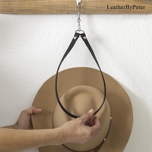 May include: A black leather hat hanger suspended from a silver-colored hook. The hanger is holding a tan-colored hat. The text "LeatherByPeter" is visible on a wooden plank above the hanger.