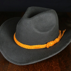 K&ouml;nnte beinhalten: Ein grauer Filzhut im Cowboy-Stil mit breiter Krempe und einem geflochtenen orangefarbenen Lederband.