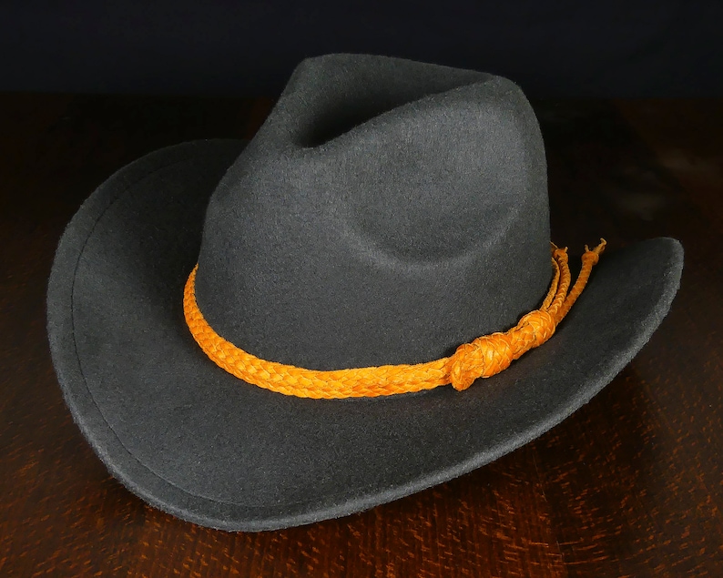 K&ouml;nnte beinhalten: Ein grauer Filzhut im Cowboy-Stil mit breiter Krempe und einem geflochtenen orangefarbenen Lederband.