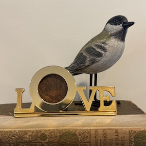 May include: A gold-toned metal sign spelling the word LOVE with a circular frame replacing the letter O, positioned in front of a small, realistic figurine of a black, white, and gray bird standing on thin black metal legs.