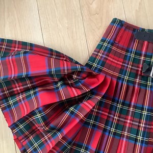Puede incluir: Una falda escocesa plisada con un patrón de cuadros tartán en rojo, verde, azul y amarillo. La prenda incluye dos correas de cuero negro con hebillas metálicas plateadas en el lateral para un ajuste regulable, mostrando pliegues profundos.