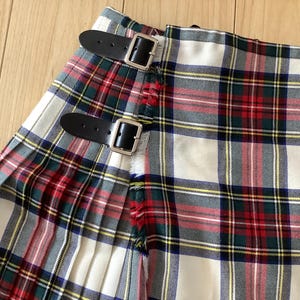Puede incluir: Una falda tipo kilt plisada con un patrón de tartán tradicional en crema, rojo, verde y azul marino. La prenda incluye dos correas de cuero negro ajustables con hebillas de metal plateado en el lateral y un detalle de borde deshilachado.