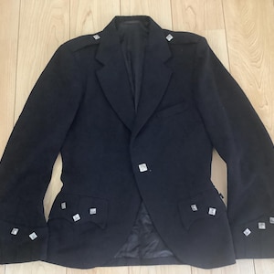 May include: A black jacket featuring a notched lapel, shoulder epaulettes, and two front pockets with flaps. The garment is adorned with small, square, metallic silver buttons on the cuffs, pocket flaps, and shoulders, and includes a single front button.