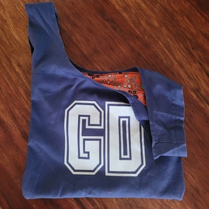 Navy blue bag with white letting in outlined capitals reading, GD. Bag is laid out flat on wooden surface. Right strap of bag is folded down to show orange print fabric lining on inside.