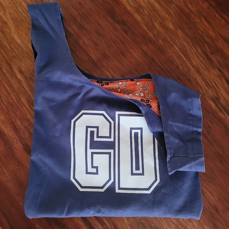 Navy blue bag with white letting in outlined capitals reading, GD. Bag is laid out flat on wooden surface. Right strap of bag is folded down to show orange print fabric lining on inside.