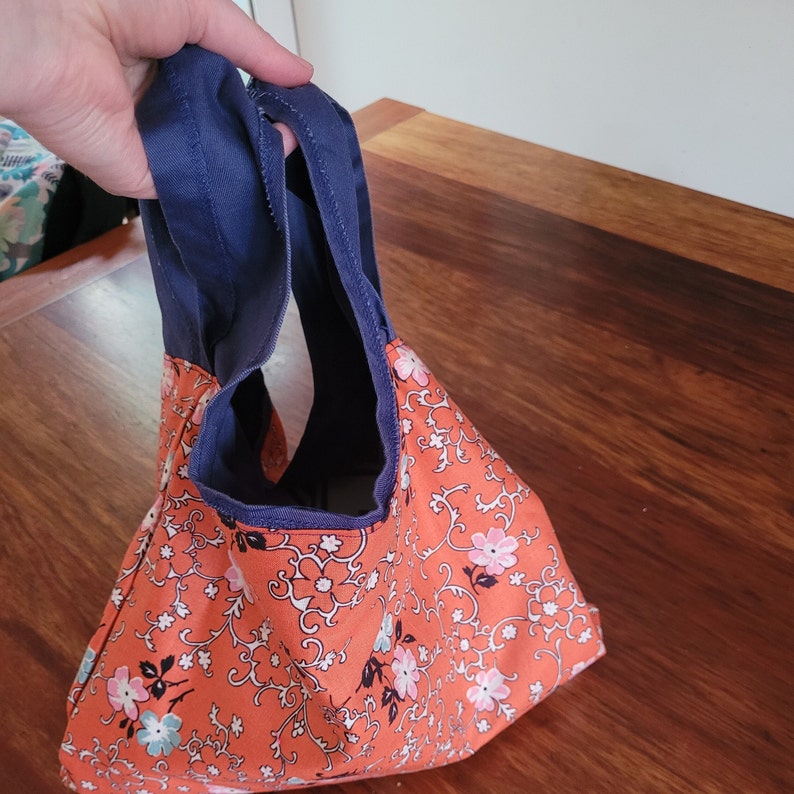 Netball bib bag reversed with orange floral lining on outside.  A hand is holding the bag by its navy blue straps.