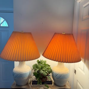 May include: A pair of table lamps featuring textured off-white ceramic bases with a light blue horizontal band of raised diagonal ridges. Each lamp is topped with a pleated, conical lampshade that emits a warm, orange-toned glow when illuminated.
