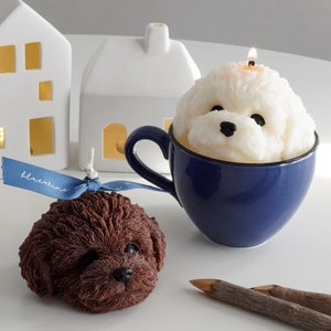 chocolate poodle soy pillar candle with blue bluewine ribbon, a lit white bichon soy pillar candle in a blue mug, wood color pencils, and two house lamps on the white round table
