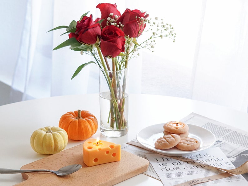 a lit yellow cheese candle and a spoon on ikea mini wood board, cookie candles on white ceramic dish, magazine papers, yellow and orange pumpkins, red roses and gypsophila in a clear vase on white round table