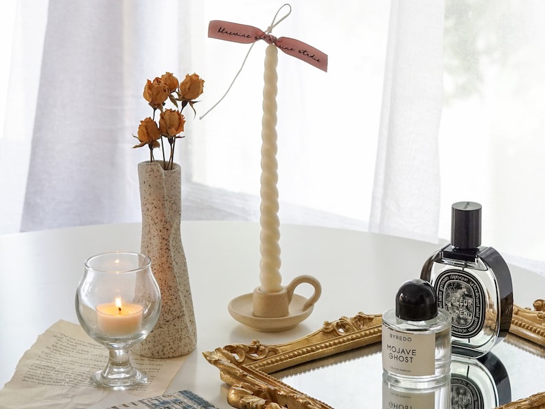 white twisted swirl soy pillar taper candle in a dotted beige ceramic candle holder, yellow rose dried flowers in unique vase and a lit tealight candle and Byredo Mojave Ghost and Diptyque Fleur de Peau placed on rectangular gold french mirror tray