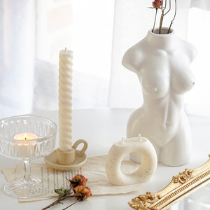 white twisted swirl taper candle in a ceramic candle holder, orange rose dried flowers, ring shape dotted candle, a lit tealight in a coupe glass, rectangular gold french mirror tray, a feminine body shape white ceramic vase on white round table