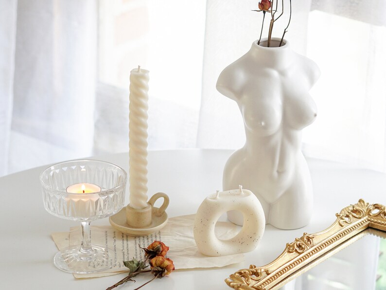 white twisted swirl taper candle in a ceramic candle holder, orange rose dried flowers, ring shape dotted candle, a lit tealight in a coupe glass, rectangular gold french mirror tray, a feminine body shape white ceramic vase on white round table