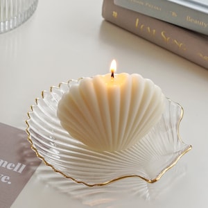 a lit white seashell minimal aesthetic soy pillar candle on a gold rim clear shell tray, two books, you smell like love beige postcard